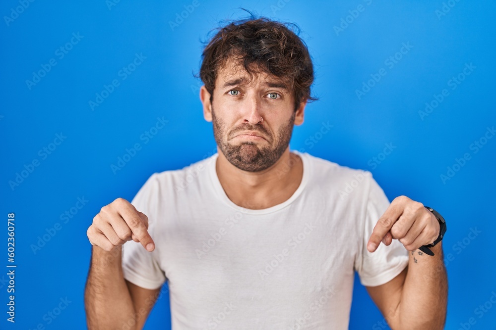 Hispanic young man standing over blue background pointing down looking sad and upset, indicating direction with fingers, unhappy and depressed.