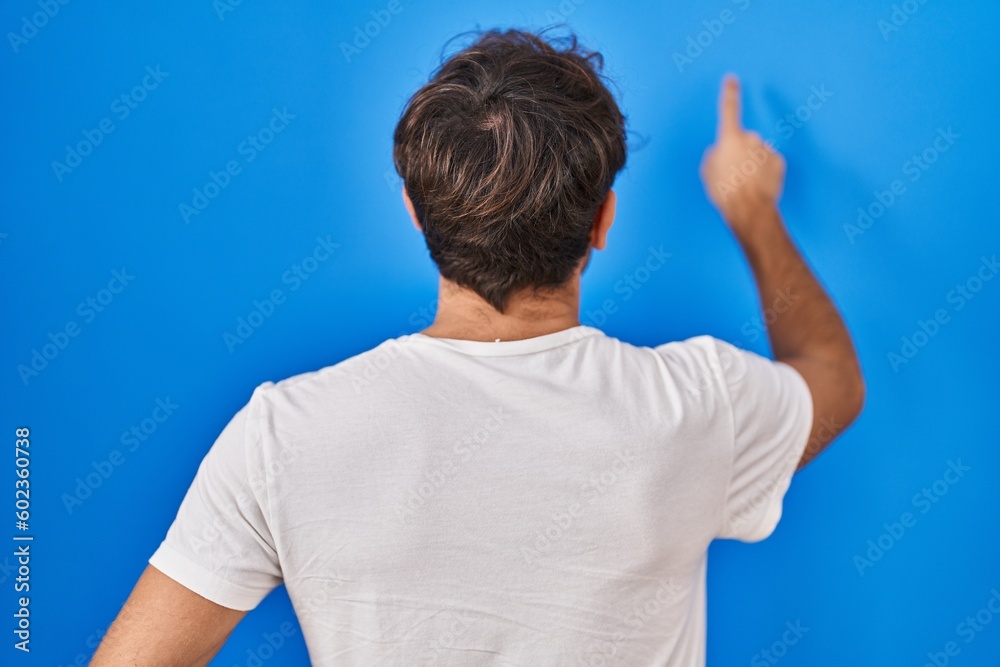 Hispanic young man standing over blue background posing backwards pointing ahead with finger hand Hispanic young man standing over blue background posing backwards pointing ahead with finger hand