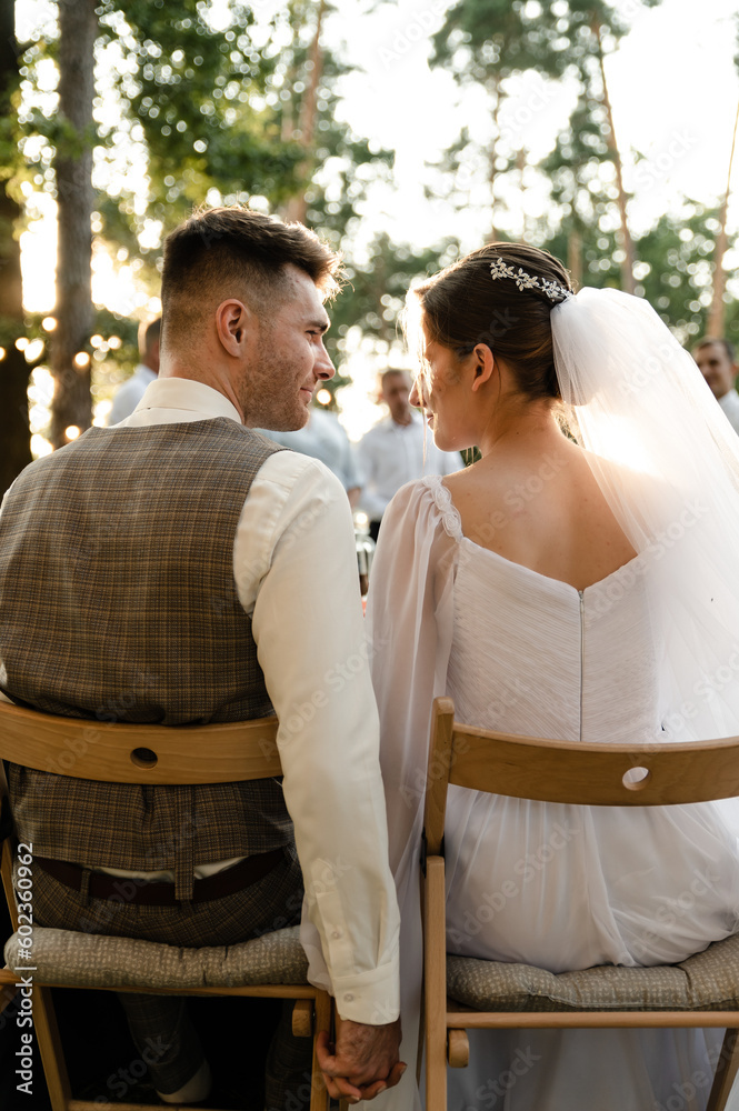 A young groom in a gray suit and a smiling brunette bride are sitting on chairs with their backs to the table, looking at each other and laughing against the background of light bulbs, garlands..
