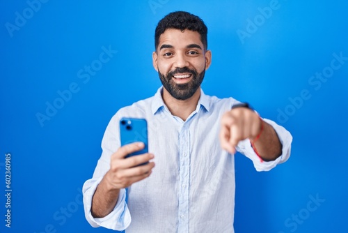 Hispanic man with beard using smartphone typing message pointing to you and the camera with fingers, smiling positive and cheerful © Krakenimages.com