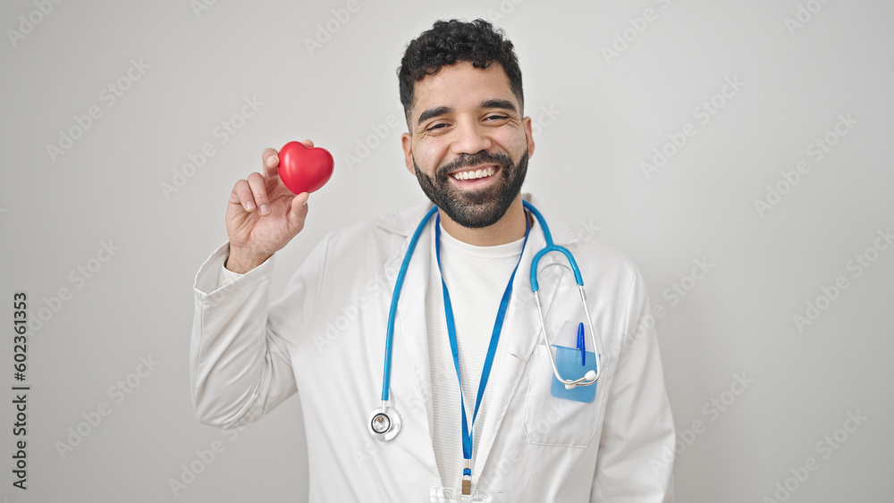 Young hispanic man doctor smiling holding heart over isolated white background Young hispanic man doctor smiling holding heart over isolated white background