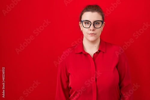 Young hispanic woman with red hair standing over red background relaxed with serious expression on face. simple and natural looking at the camera.