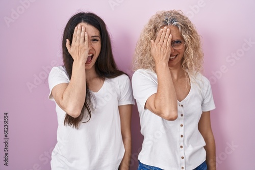 Mother and daughter standing together over pink background covering one eye with hand, confident smile on face and surprise emotion.