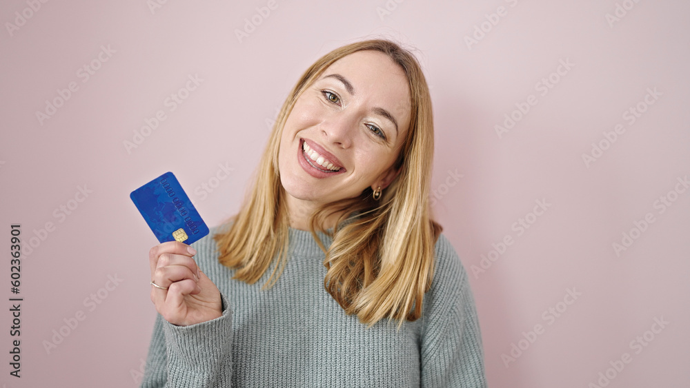 Young blonde woman smiling confident holding credit card over isolated pink background Young blonde woman smiling confident holding credit card over isolated pink background