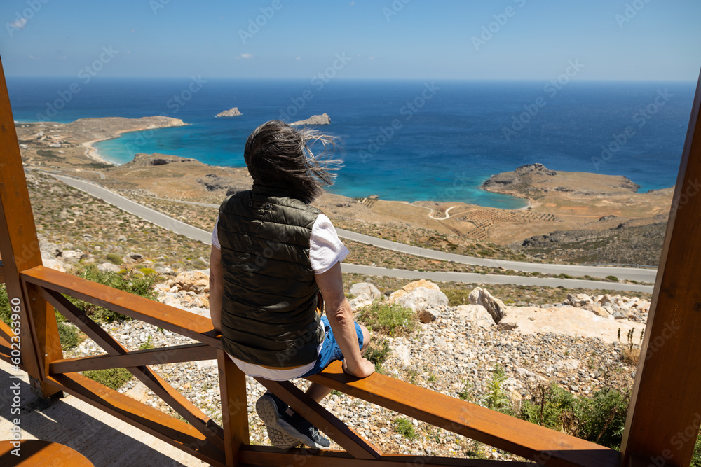 Long-haired man sitting on a fence and looking at the beautiful coast of Crete, Greece Long-haired man sitting on a fence and looking at the beautiful coast of Crete, Greece