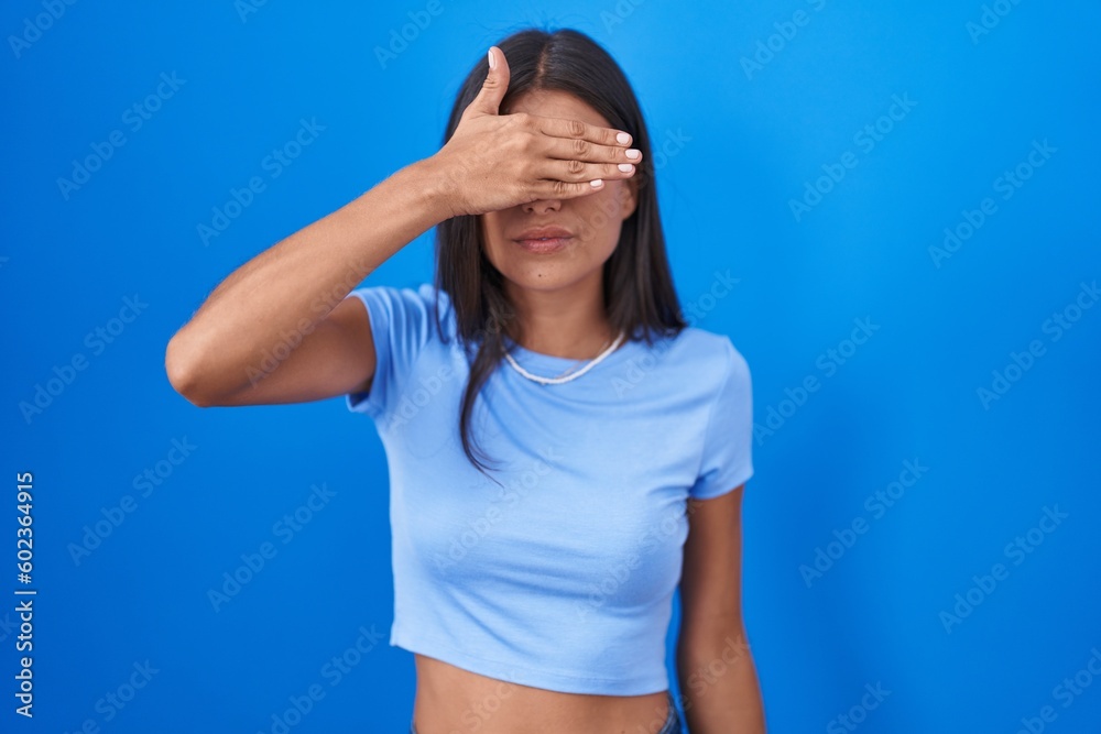Brunette young woman standing over blue background covering eyes with hand, looking serious and sad. sightless, hiding and rejection concept Brunette young woman standing over blue background covering eyes with hand, looking serious and sad. sightless, hiding and rejection concept