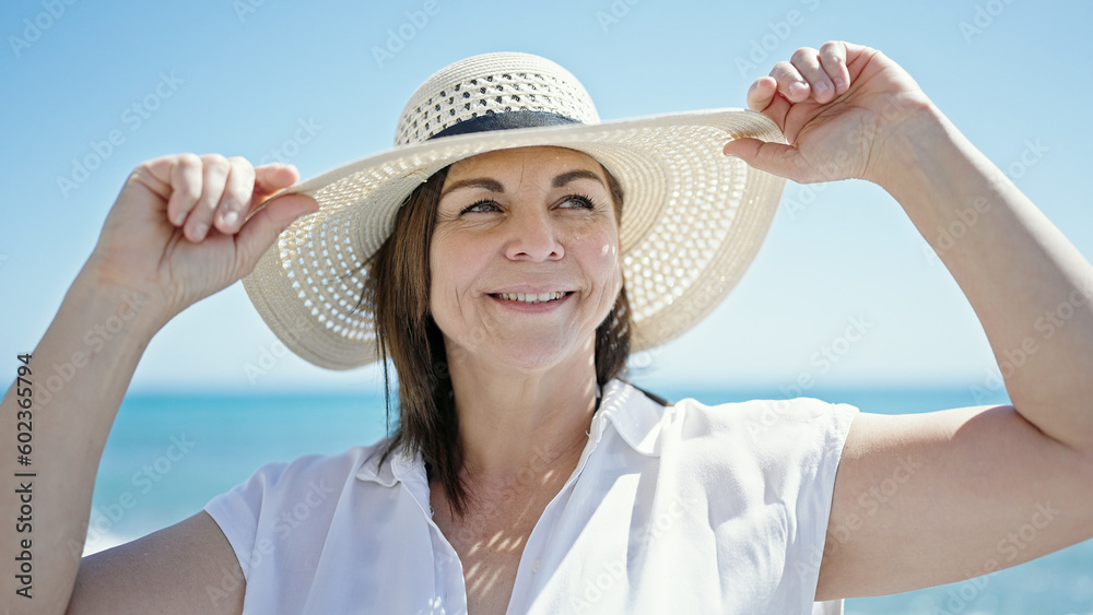 Middle age hispanic woman tourist smiling holding summer hat at the beach