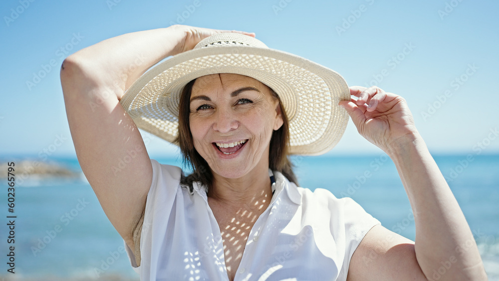 Middle age hispanic woman tourist smiling holding summer hat at the beach