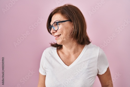 Middle age hispanic woman standing over pink background looking away to side with smile on face, natural expression. laughing confident.