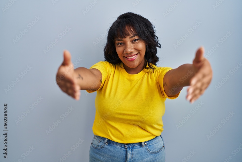 Hispanic woman standing over blue background looking at the camera smiling with open arms for hug. cheerful expression embracing happiness. Hispanic woman standing over blue background looking at the camera smiling with open arms for hug. cheerful expression embracing happiness.