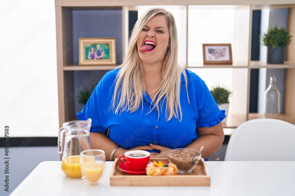 Caucasian plus size woman eating breakfast at home sticking tongue out happy with funny expression. emotion concept.