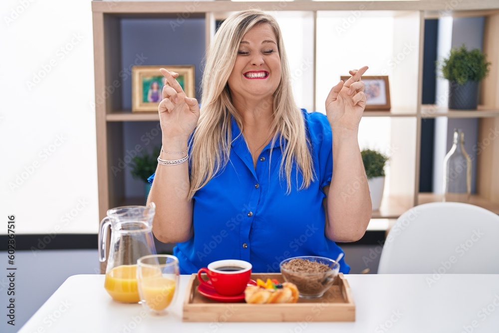 Caucasian plus size woman eating breakfast at home gesturing finger crossed smiling with hope and eyes closed. luck and superstitious concept.