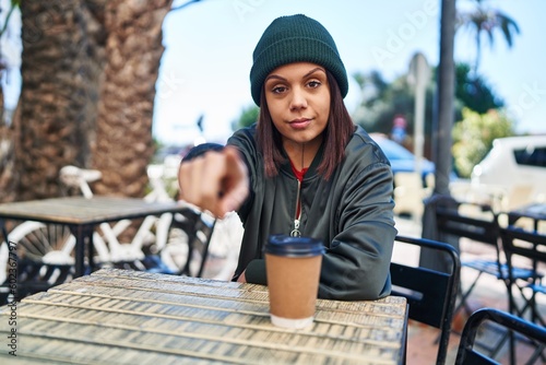 Young hispanic woman drinking a cup of coffee outdoors pointing with finger to the camera and to you, confident gesture looking serious