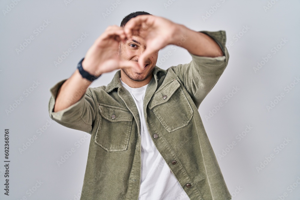 Young hispanic man standing over isolated background smiling in love doing heart symbol shape with hands. romantic concept. Young hispanic man standing over isolated background smiling in love doing heart symbol shape with hands. romantic concept.
