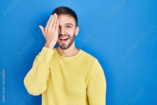 Hispanic man standing over blue background covering one eye with hand, confident smile on face and surprise emotion.
