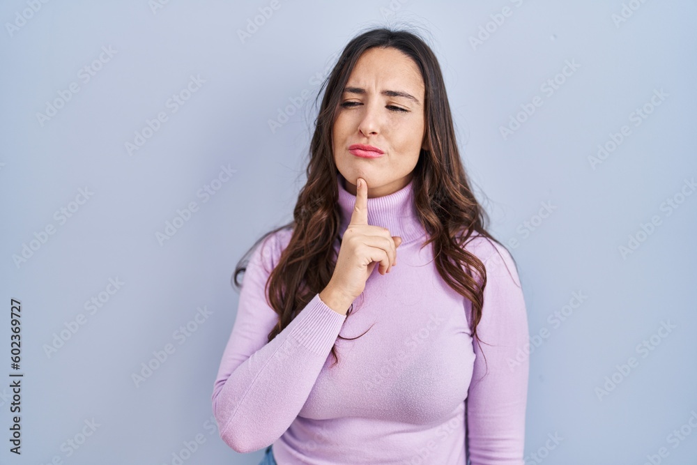 Young brunette woman standing over blue background thinking concentrated about doubt with finger on chin and looking up wondering