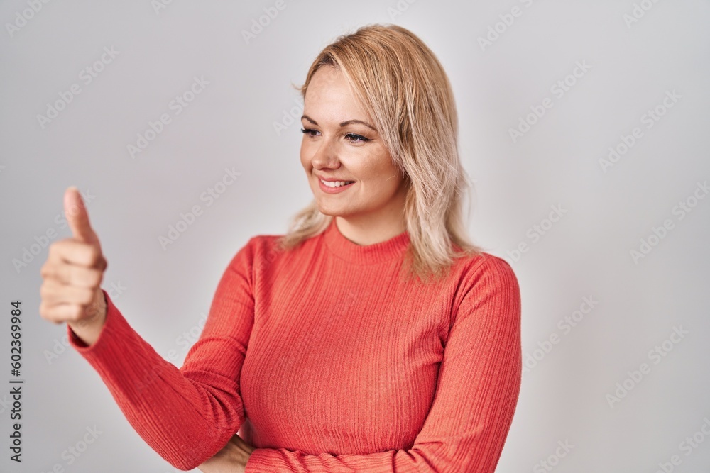Fototapeta premium Blonde woman standing over isolated background looking proud, smiling doing thumbs up gesture to the side