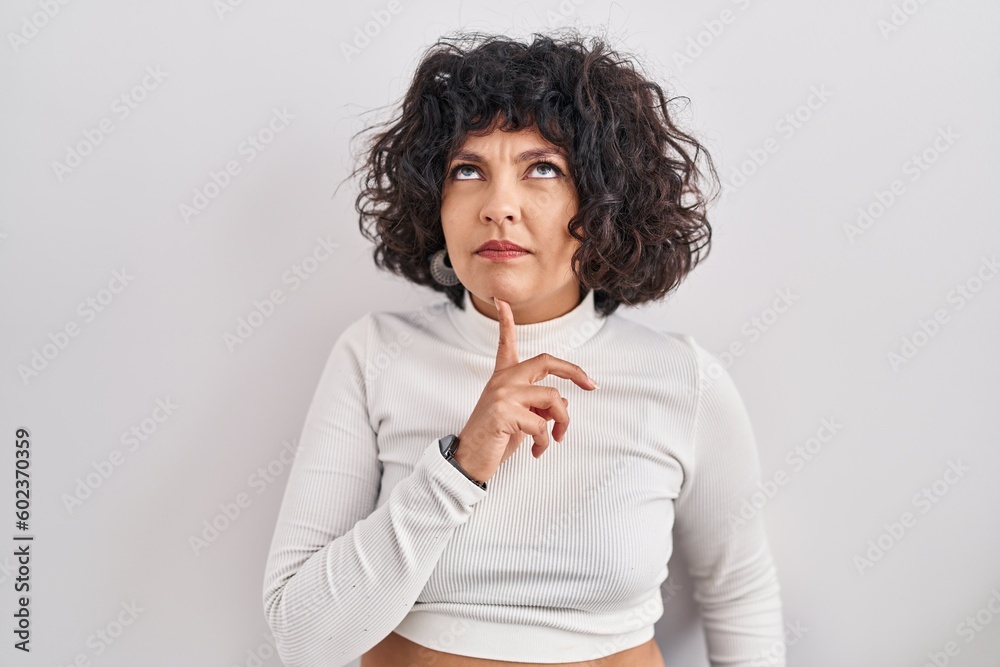 Hispanic woman with curly hair standing over isolated background thinking concentrated about doubt with finger on chin and looking up wondering Hispanic woman with curly hair standing over isolated background thinking concentrated about doubt with finger on chin and looking up wondering