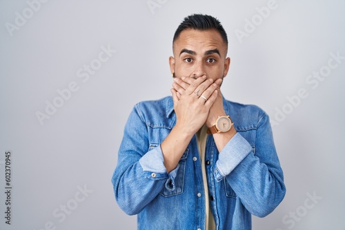 Young hispanic man standing over isolated background shocked covering mouth with hands for mistake. secret concept.