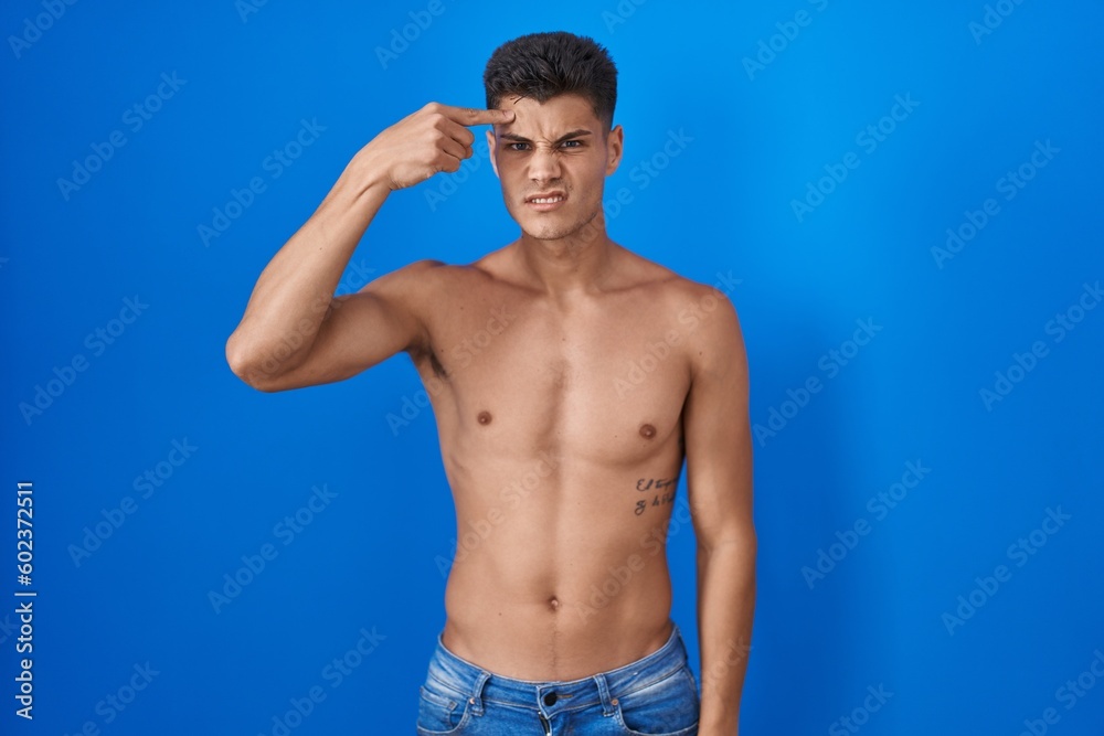 Young hispanic man standing shirtless over blue background pointing unhappy to pimple on forehead, ugly infection of blackhead. acne and skin problem