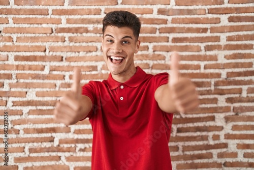Young hispanic man standing over bricks wall approving doing positive gesture with hand, thumbs up smiling and happy for success. winner gesture.