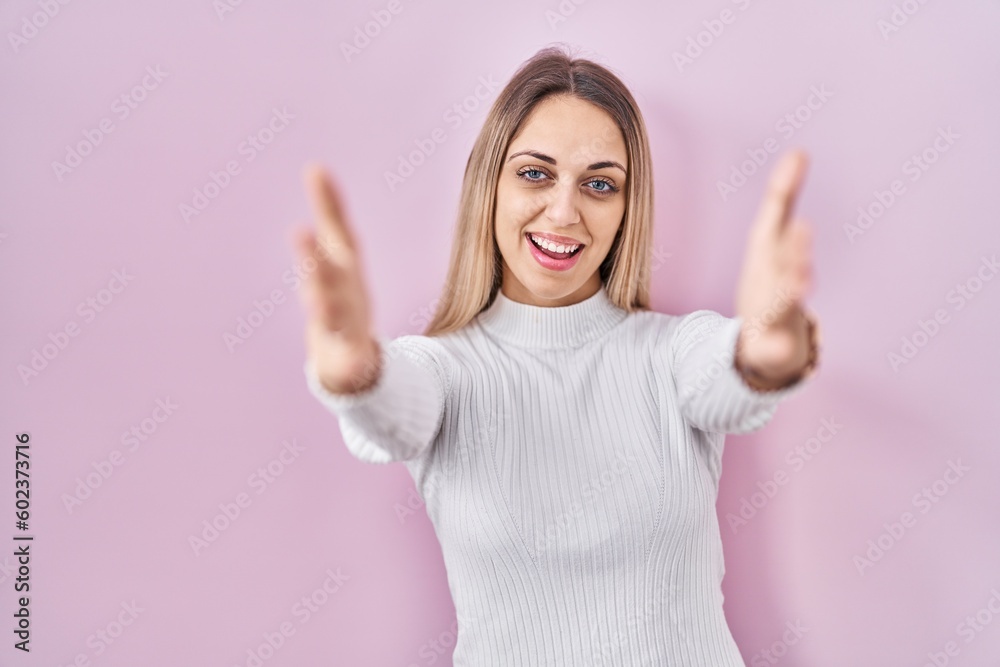 Young blonde woman wearing white sweater over pink background looking at the camera smiling with open arms for hug. cheerful expression embracing happiness. Young blonde woman wearing white sweater over pink background looking at the camera smiling with open arms for hug. cheerful expression embracing happiness.
