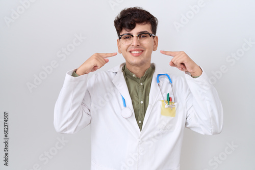 Young non binary man wearing doctor uniform and stethoscope smiling cheerful showing and pointing with fingers teeth and mouth. dental health concept.