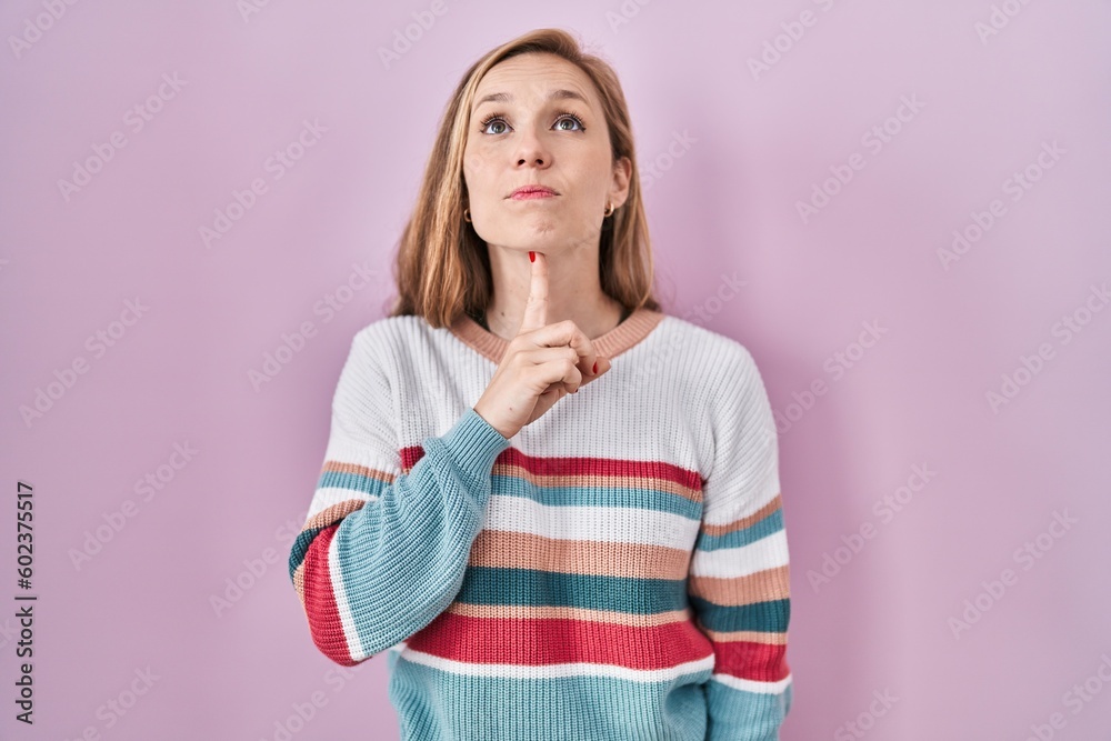 Young blonde woman standing over pink background thinking concentrated about doubt with finger on chin and looking up wondering Young blonde woman standing over pink background thinking concentrated about doubt with finger on chin and looking up wondering