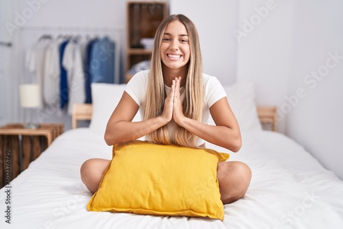 Young blonde woman sitting on the bed with pillow at home praying with hands together asking for forgiveness smiling confident.
