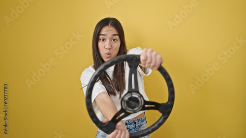 Young beautiful hispanic woman using steering wheel as a driver over isolated yellow background
