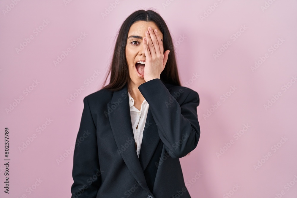 Young brunette woman wearing business style over pink background covering one eye with hand, confident smile on face and surprise emotion. Young brunette woman wearing business style over pink background covering one eye with hand, confident smile on face and surprise emotion.