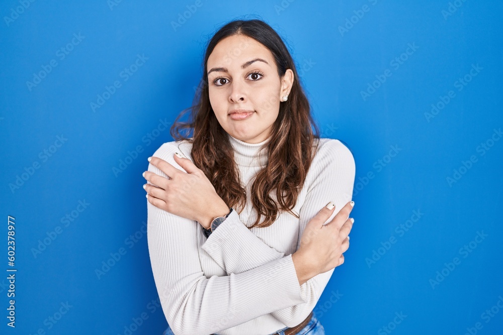 Fototapeta premium Young hispanic woman standing over blue background shaking and freezing for winter cold with sad and shock expression on face