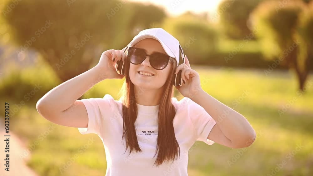 Young happy woman in a white cap, sunglasses listens to music in wireless headphones while walking in the park,sunny day Young happy woman in a white cap, sunglasses listens to music in wireless headphones while walking in the park,sunny day