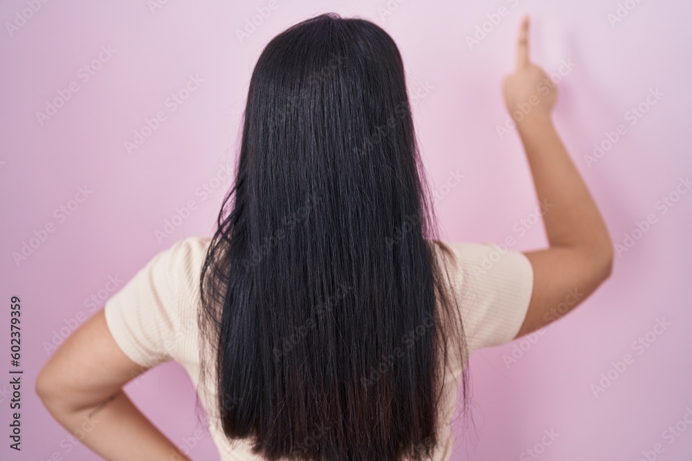 Fototapeta premium Young hispanic woman standing over pink background posing backwards pointing ahead with finger hand