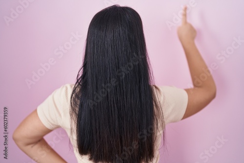 Young hispanic woman standing over pink background posing backwards pointing ahead with finger hand