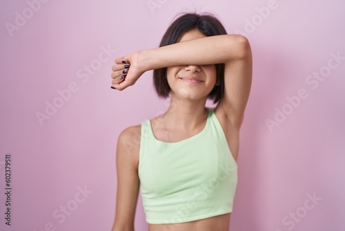 Young girl standing over pink background covering eyes with arm smiling cheerful and funny. blind concept.