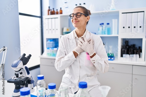 Young woman working at scientist laboratory smiling with hands on chest, eyes closed with grateful gesture on face. health concept.