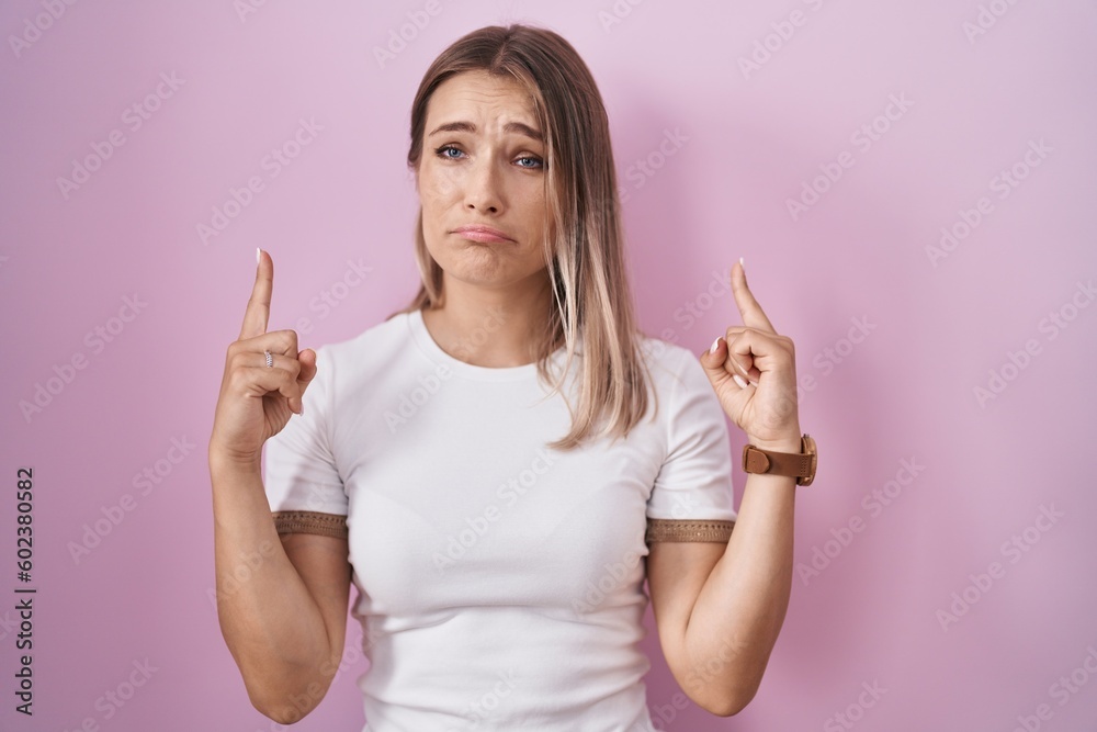 Blonde caucasian woman standing over pink background pointing up looking sad and upset, indicating direction with fingers, unhappy and depressed. Blonde caucasian woman standing over pink background pointing up looking sad and upset, indicating direction with fingers, unhappy and depressed.
