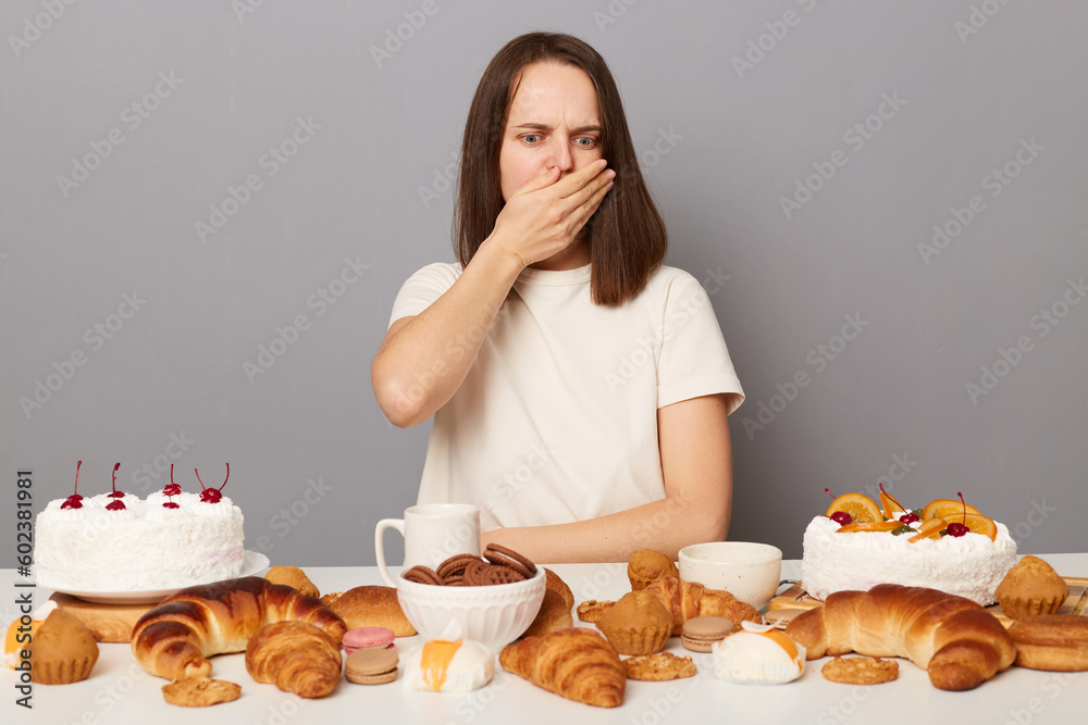 Sick woman wearing white T-shirt sitting among sugary products and various desserts, isolated over gray background, being overeating, covering mouth with palm, felling nausea. Sick woman wearing white T-shirt sitting among sugary products and various desserts, isolated over gray background, being overeating, covering mouth with palm, felling nausea.