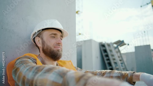 Young foreman sitting outdoors and feeling tired after overwork. Caucasian worker in reflective vest and helmet at construction site. Slow motion