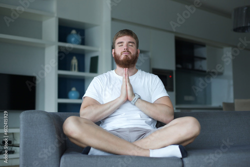 Young man with a beard alone at home, relax in living room ,sitting on the couch and meditating in the lotus position, in casual clothes