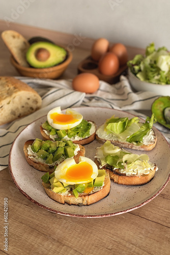 Ciabatta slices with cream cheese, avocado pieces and boiled egg on a ceramic plate. Step 7 of the healthy toast recipe with avocado, cream cheese, boiled egg and green salad. Healthy lifestyle.
