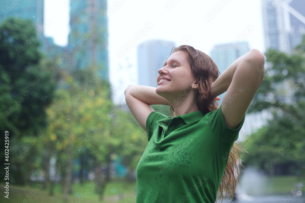 Positive young woman smiling during the rain in the park. Cheerful female enjoying the rain outdoors. Woman looking up and catching the rain drop with hands. Breath deep and relax