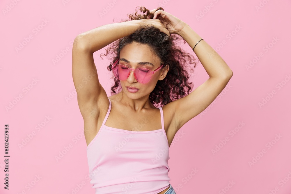 Happy woman hands up waxing her armpits, shaving, with curly hair in a pink tank top and jeans on a pink background wearing sunglasses with a nice tan, copy space Happy woman hands up waxing her armpits, shaving, with curly hair in a pink tank top and jeans on a pink background wearing sunglasses with a nice tan, copy space