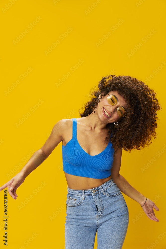 Woman with curly afro hair in a blue T-shirt on a yellow background dancing flying hair with sunglasses yellow, hand signs, look into the camera, smile with teeth and happiness, copy space Woman with curly afro hair in a blue T-shirt on a yellow background dancing flying hair with sunglasses yellow, hand signs, look into the camera, smile with teeth and happiness, copy space
