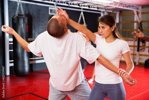 Latin woman exercising palm strike to chin with caucasian man in gym during group self-defence training. African-american man trainer standing and watching them.