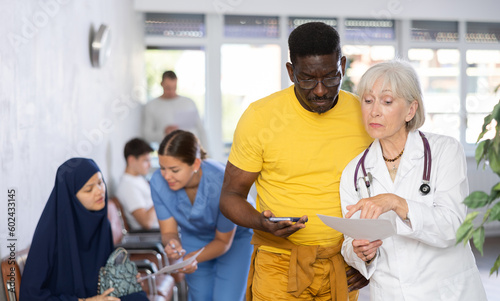 Experienced elderly female doctor explaining results of medical examination to focused african american male patient while standing in lobby of clinic