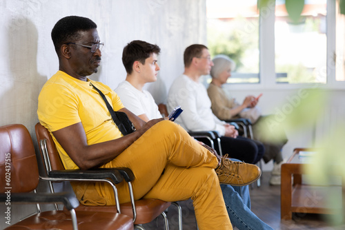 Adult African American in casual clothes sitting in lobby of medical clinic, browsing Internet while waiting for appointment with doctor