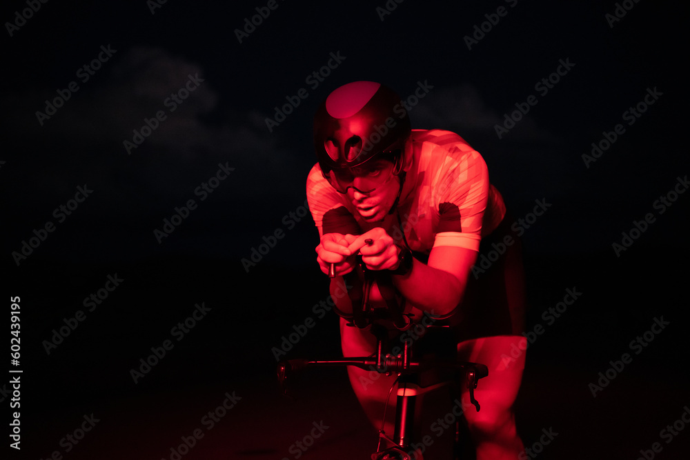 A triathlete rides his bike in the darkness of night, pushing himself to prepare for a marathon. The contrast between the darkness and the light of his bike creates a sense of drama and highlights the A triathlete rides his bike in the darkness of night, pushing himself to prepare for a marathon. The contrast between the darkness and the light of his bike creates a sense of drama and highlights the