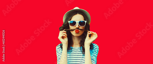 Portrait of funny young woman showing mustache her hair blowing lips with red lipstick sending air kiss on background